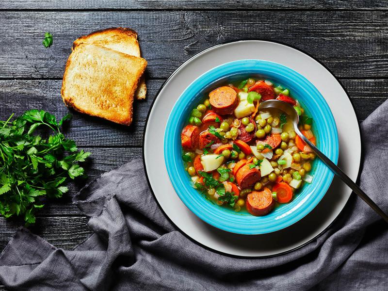 Smoked sausage soup in a bowl on a table with toast fresh herbs and table cloth.